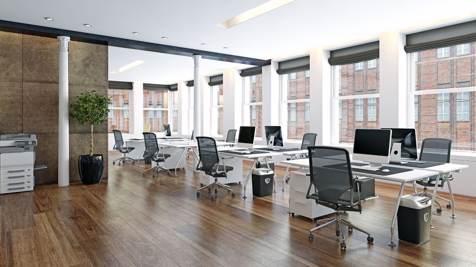 Modern open office space with white desks, black mesh chairs, and large windows overlooking brick buildings.