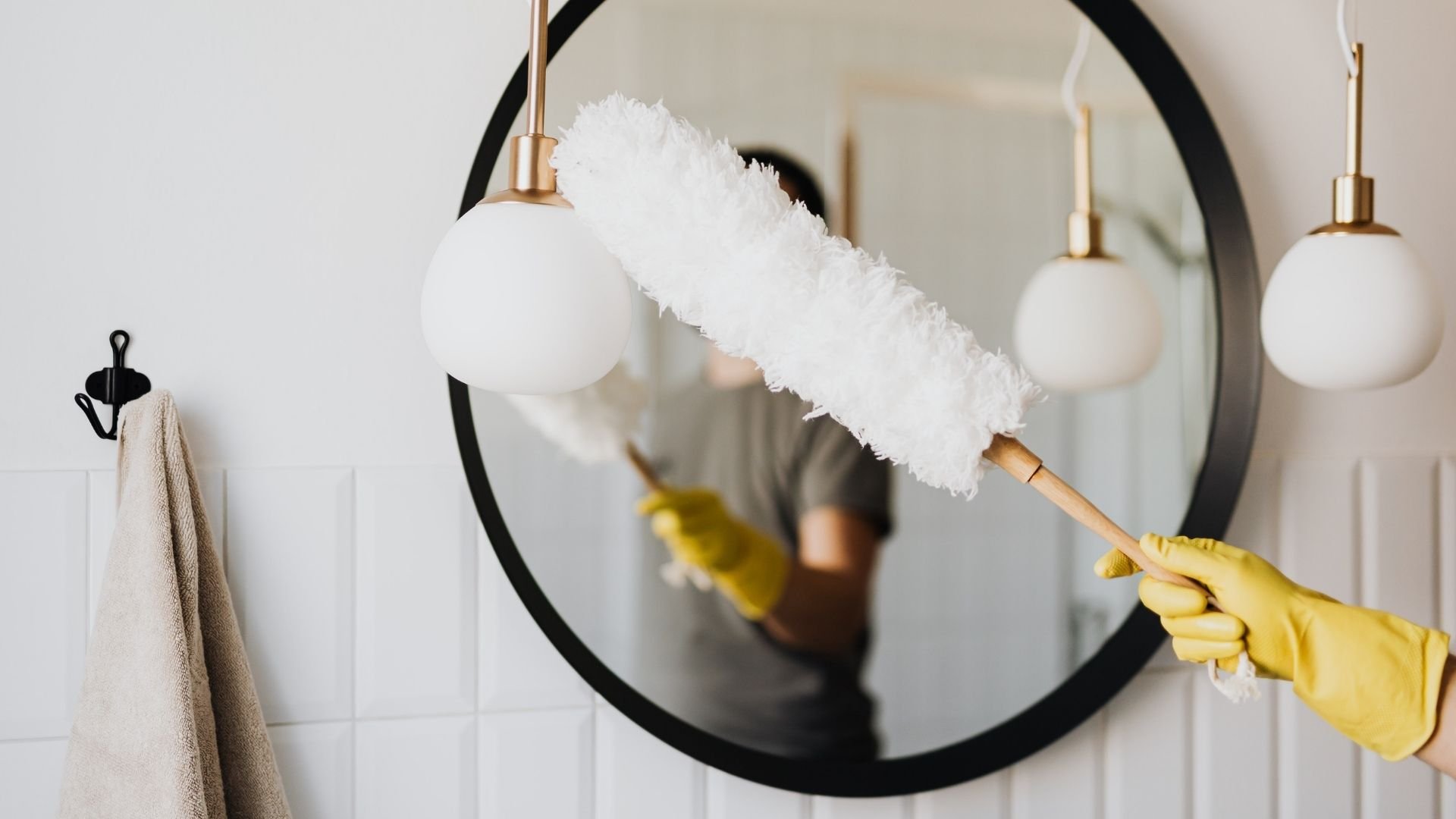 Hand in yellow glove cleaning mirror with white duster near bathroom pendants.